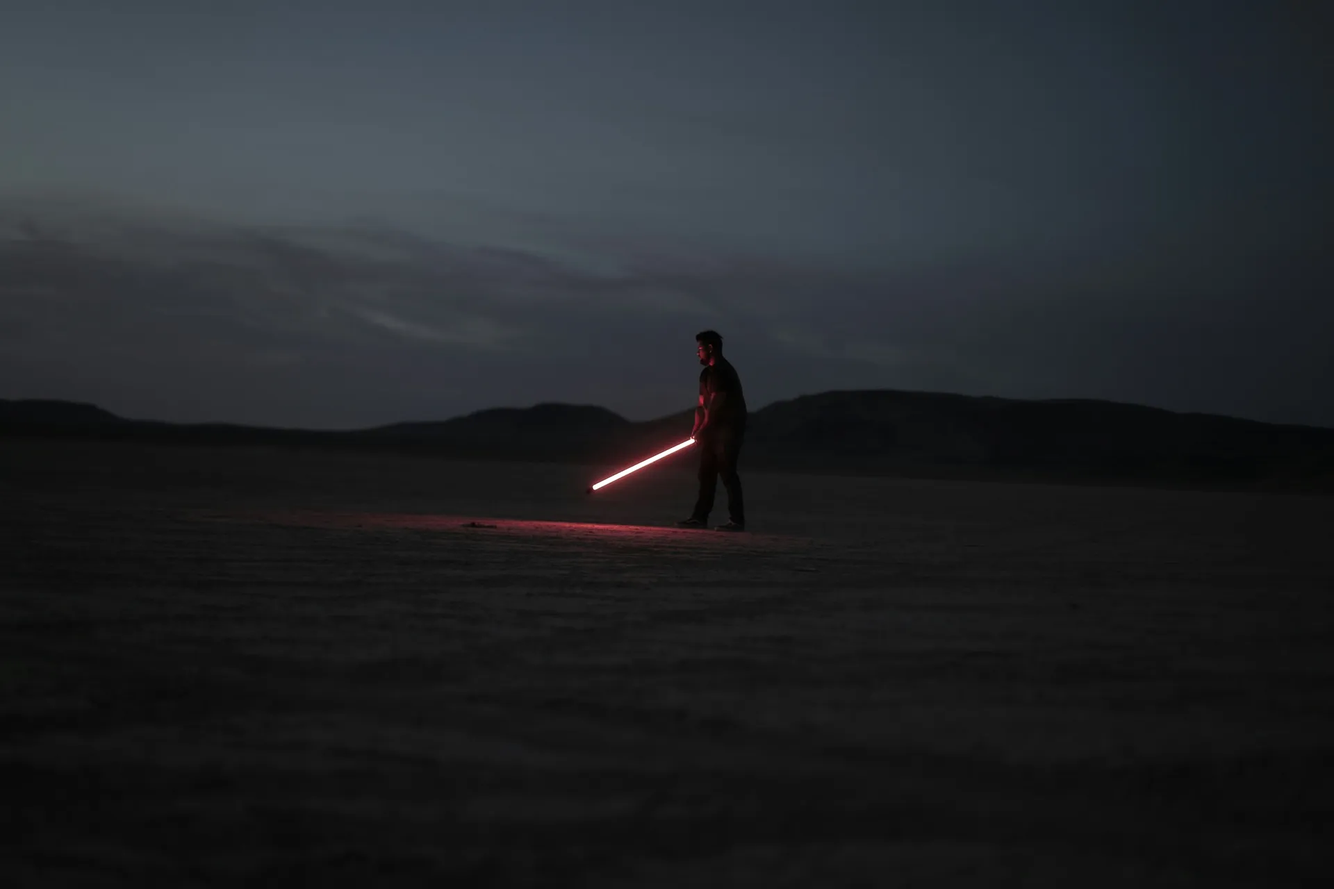A silhouetted figure holding a glowing red lightsaber in the desert at dusk