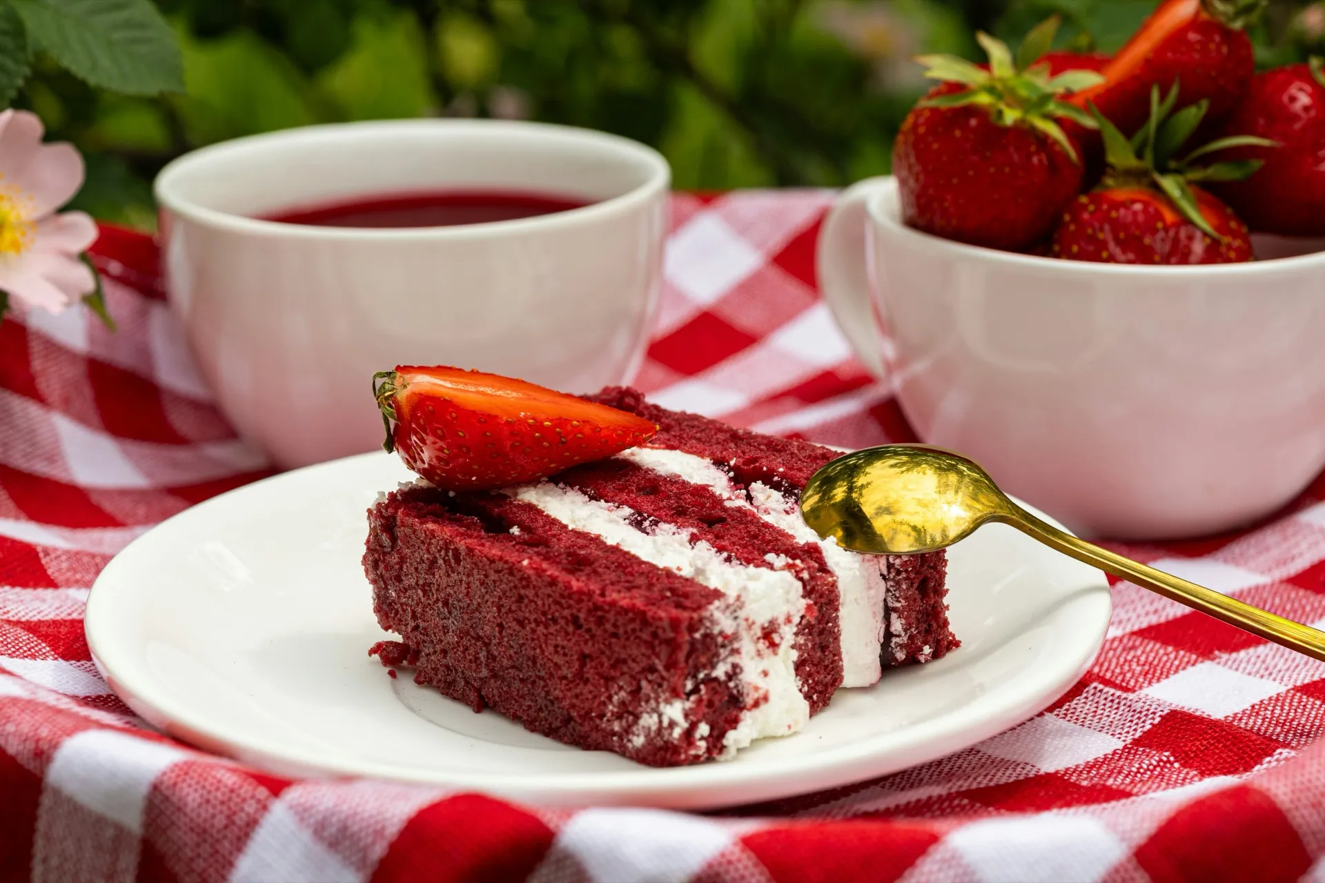 A slice of red velvet cake with a strawberry, beside cups of red tea on a red checked tablecloth