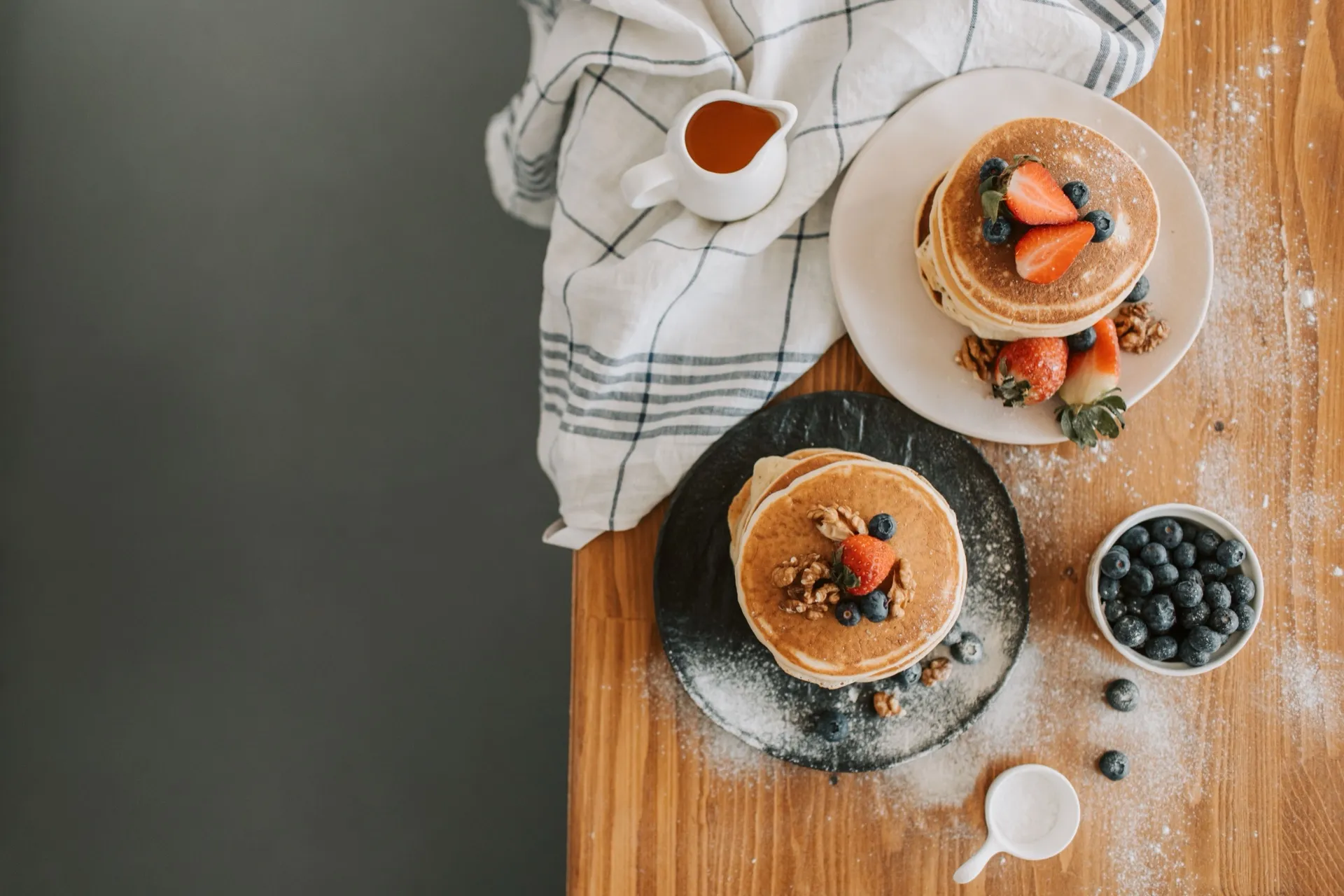 Stacks of pancakes with strawberries, blueberries and a jug of syrup on a wooden table